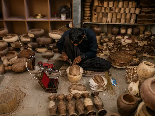 Craftsperson hand-carving a wooden vessel in a workshop filled with turned pots and tools.