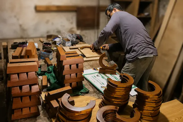 Woodworker wearing glasses shaping curved wooden pieces at a cluttered workshop table in a woodshopedition note: rings and blocks nearby