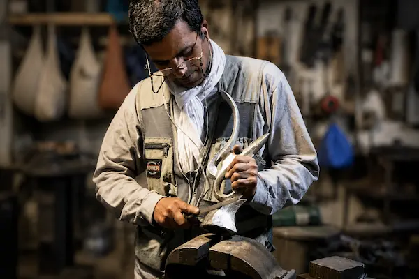 Metalworker wearing safety glasses and gloves shapes a curved metal piece on a bench vise in a workshop.
