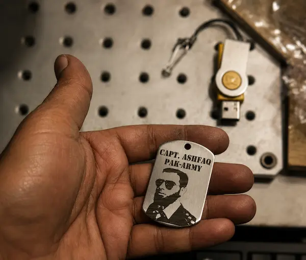 Close-up of a hand holding a small metal dog tag with a portrait and the text ‘CAPT. ? PAK ARMY’ against a perforated metal surface; a USB drive is visible in the background.