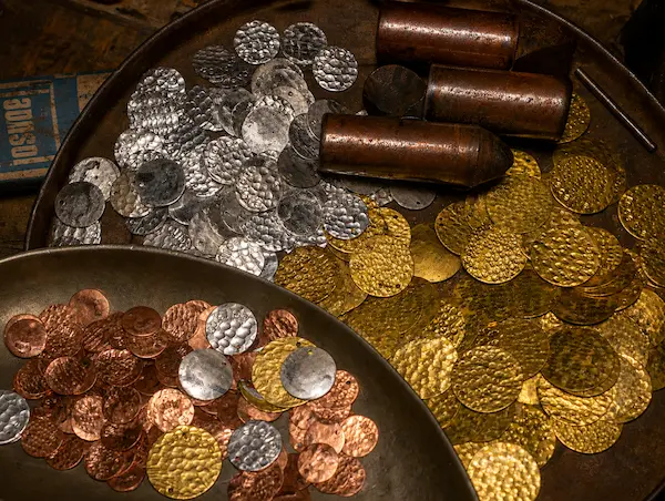 Coins in copper, silver, and gold tones scattered on metal trays; includes a few cylindrical copper cartridges nearby, suggesting a treasure hoard.