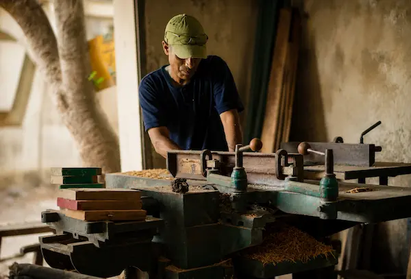 A woodworker wearing a cap operates a bench planer in a dusty workshop with wood shavings around the machine.