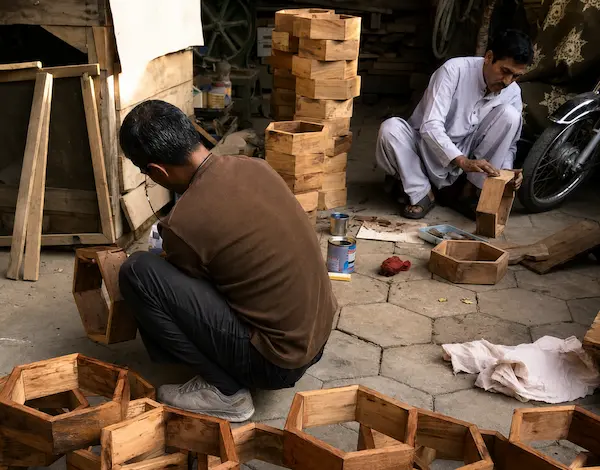 Two men work on wooden crates in a rustic workshop, kneeling on a stone floor surrounded by stacked wood blocks and tools