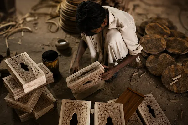 A man sits on a dusty workshop floor carving decorative stone blocks with intricate relief patterns, surrounded by carved panels and tools.